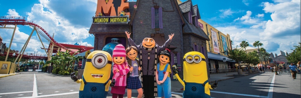 The movie characters Gru, his three smiling daughters, and two Minions pose in front of the Despicable Me Minion Mayhem attraction.